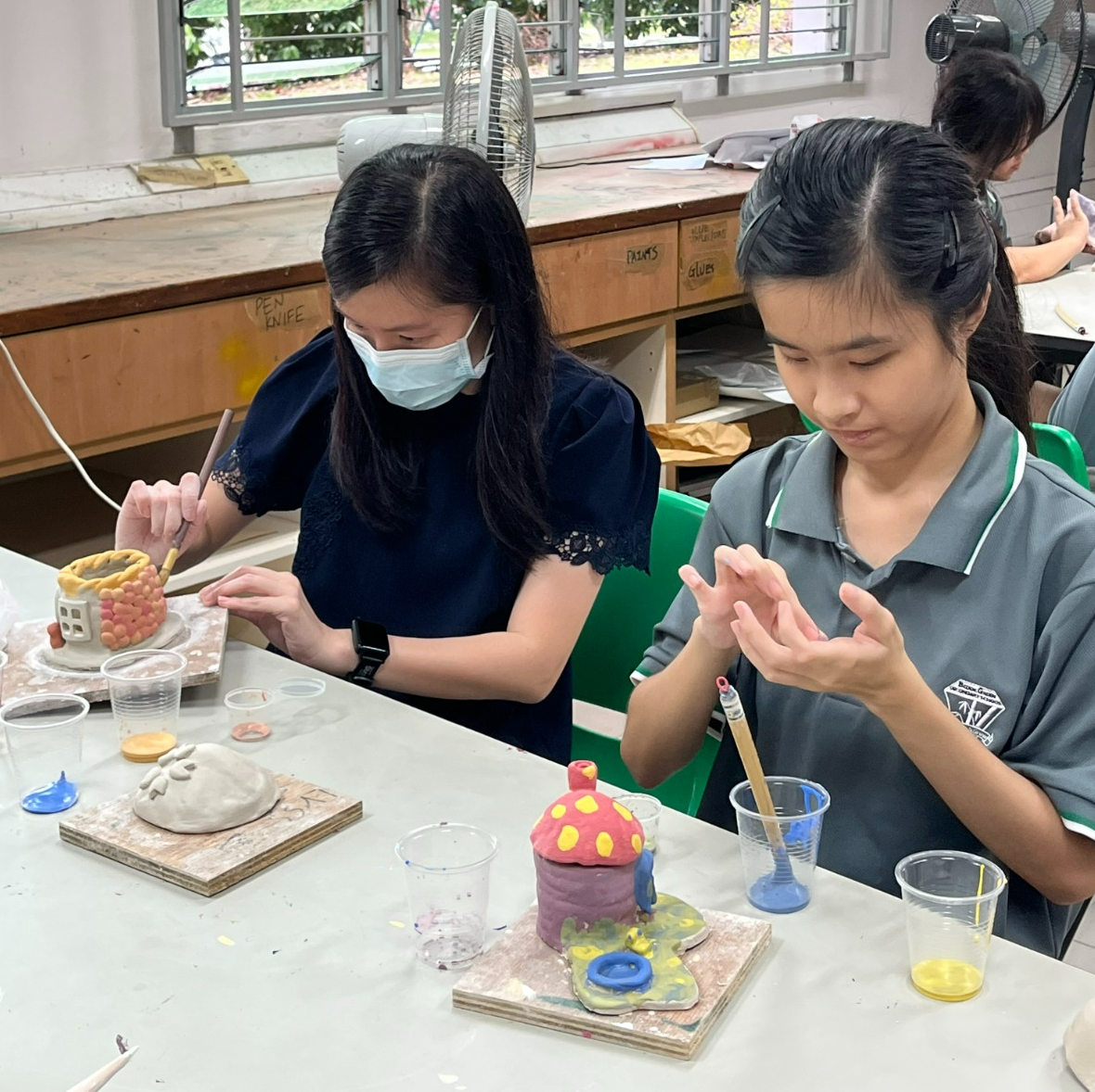 Two girls paint colorful clay sculptures at a table in an art classroom, one wearing a face mask.